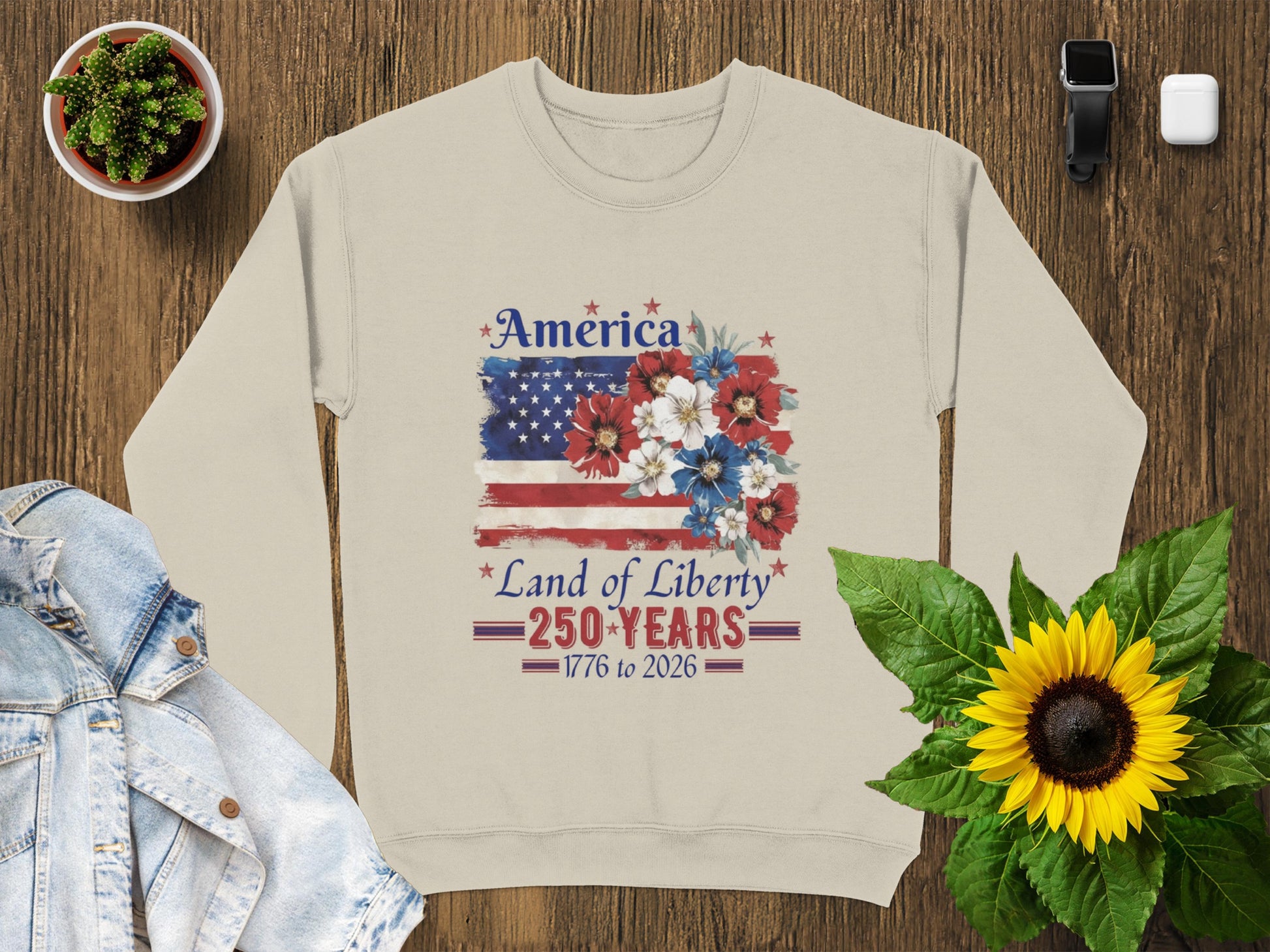 Light tan sweatshirt with "America Land of Liberty 1776-2026" text and US flag floral design.