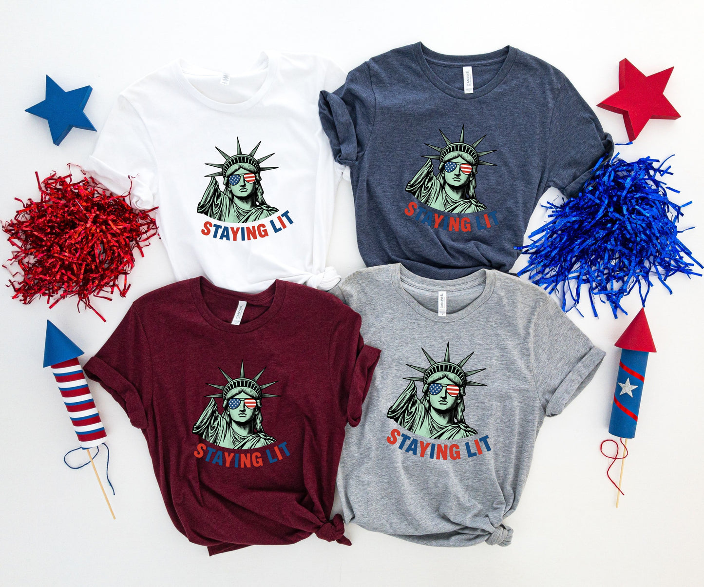 Four t-shirts with Statue of Liberty and "Staying Lit" text, arranged with fireworks.