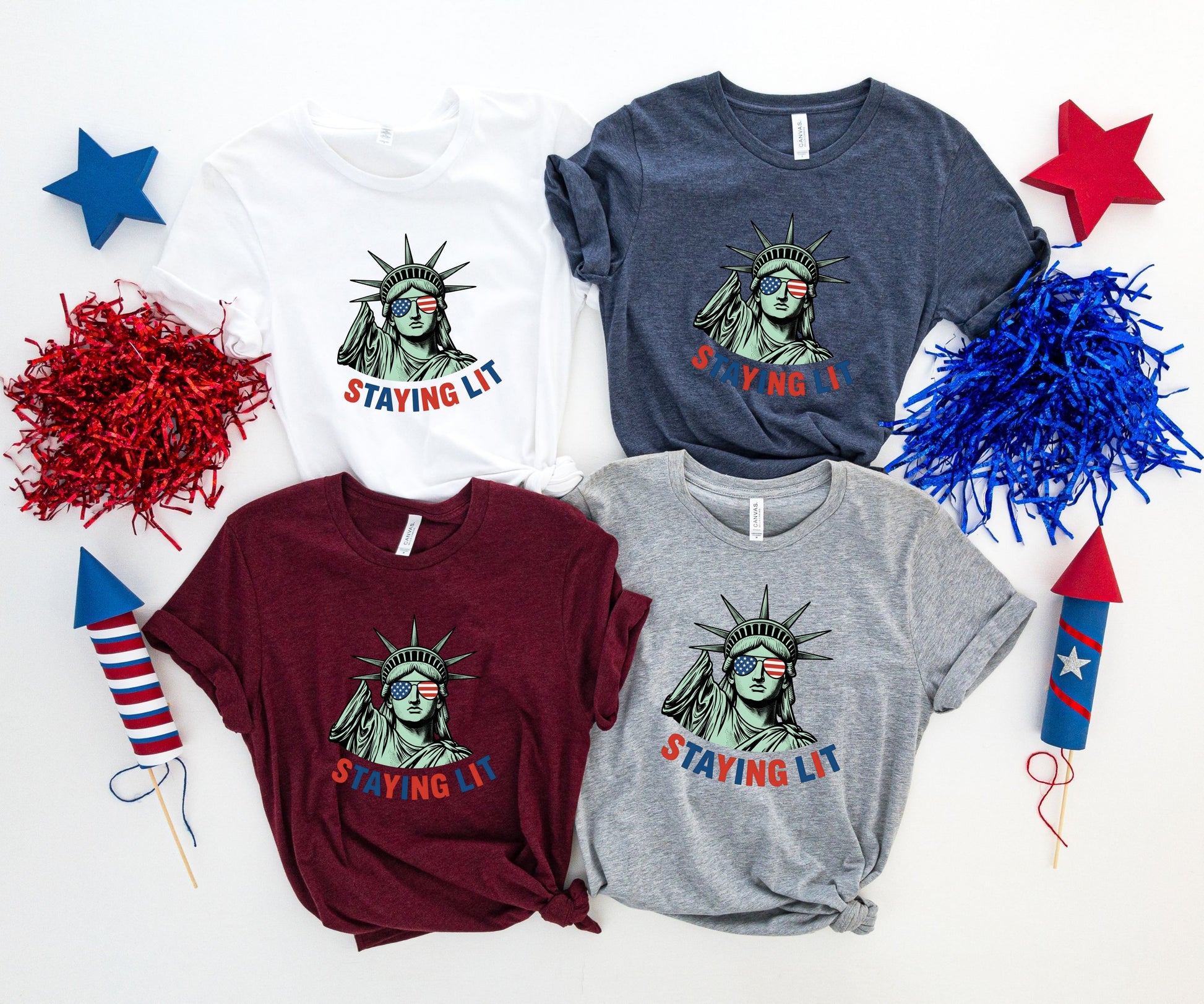 Four t-shirts with Statue of Liberty and "Staying Lit" text, arranged with fireworks.