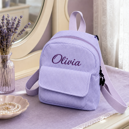 A small lavender corduroy mini backpack with the name 'Olivia' embroidered on the front, styled on a table next to a vase of dried lavender and a dish of pearls.