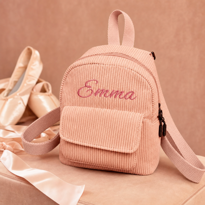 A personalized pink corduroy mini backpack with the name 'Emma' embroidered on the front, styled with ballet shoes and a ribbon on a matching pink background.