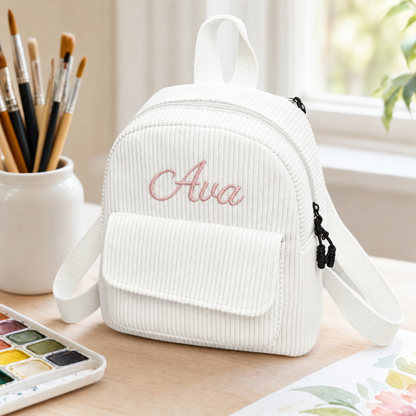 A small white corduroy mini backpack with the name 'Ava' embroidered in pink on the front pocket, shown on a wooden desk next to art supplies.