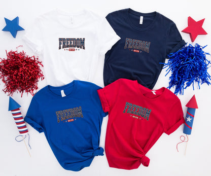 Four t-shirts with "FREEDOM EST 1776" graphic in white, navy, royal blue, and red.