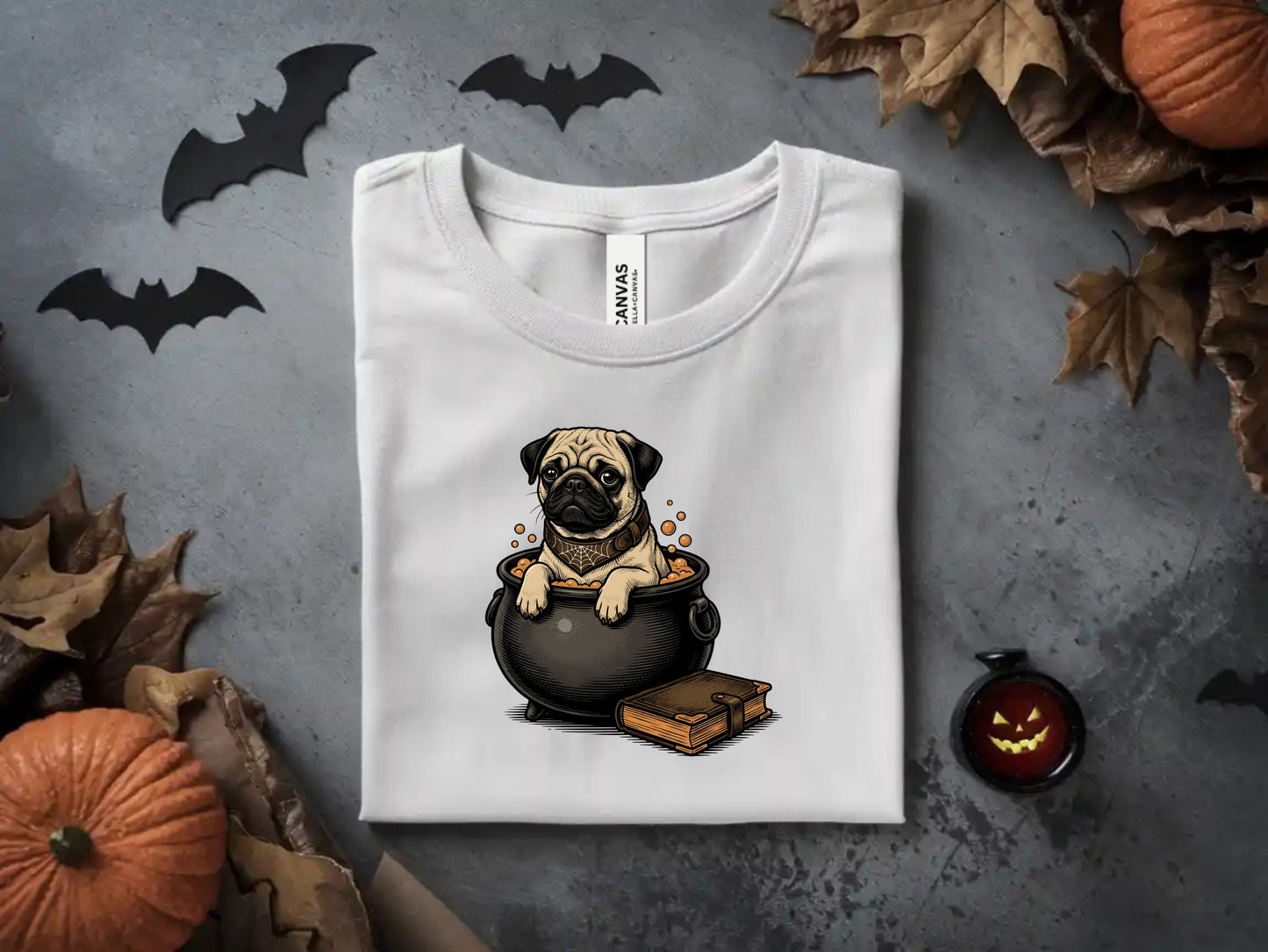 White t-shirt with a pug sitting in a cauldron, next to a book, with Halloween decorations.