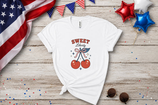 White t-shirt with "Sweet Liberty" text and cherry graphic on a wooden background.