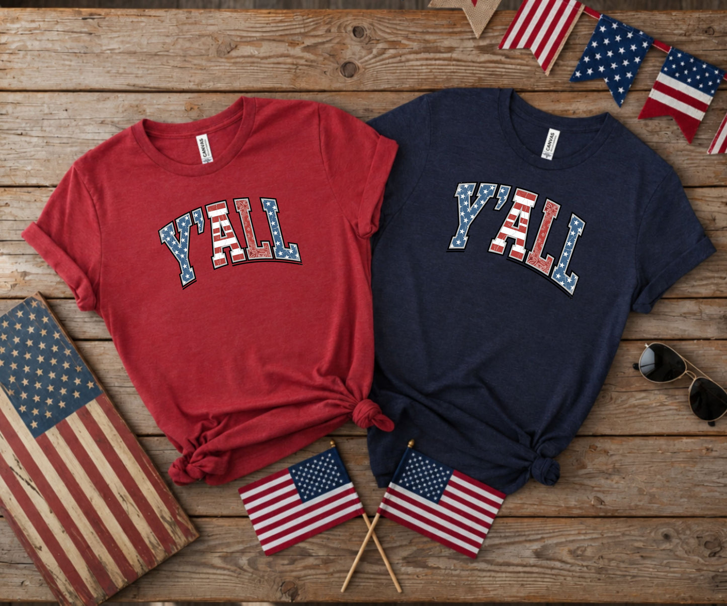 Two t-shirts with "Y'ALL" in American flag design, on wooden surface.
